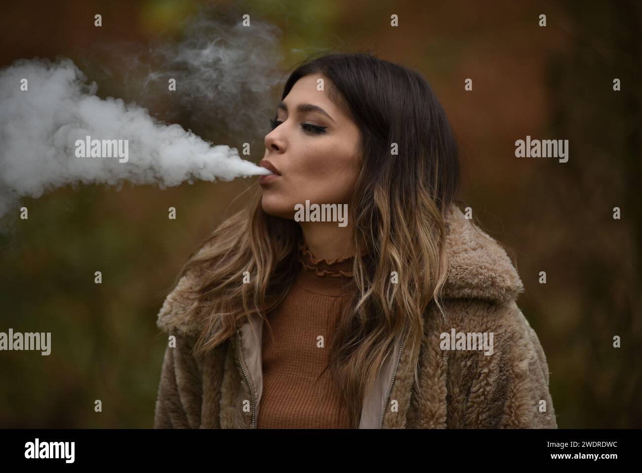 Una donna con i capelli lunghi e una giacca che fuma in un parco Foto Stock