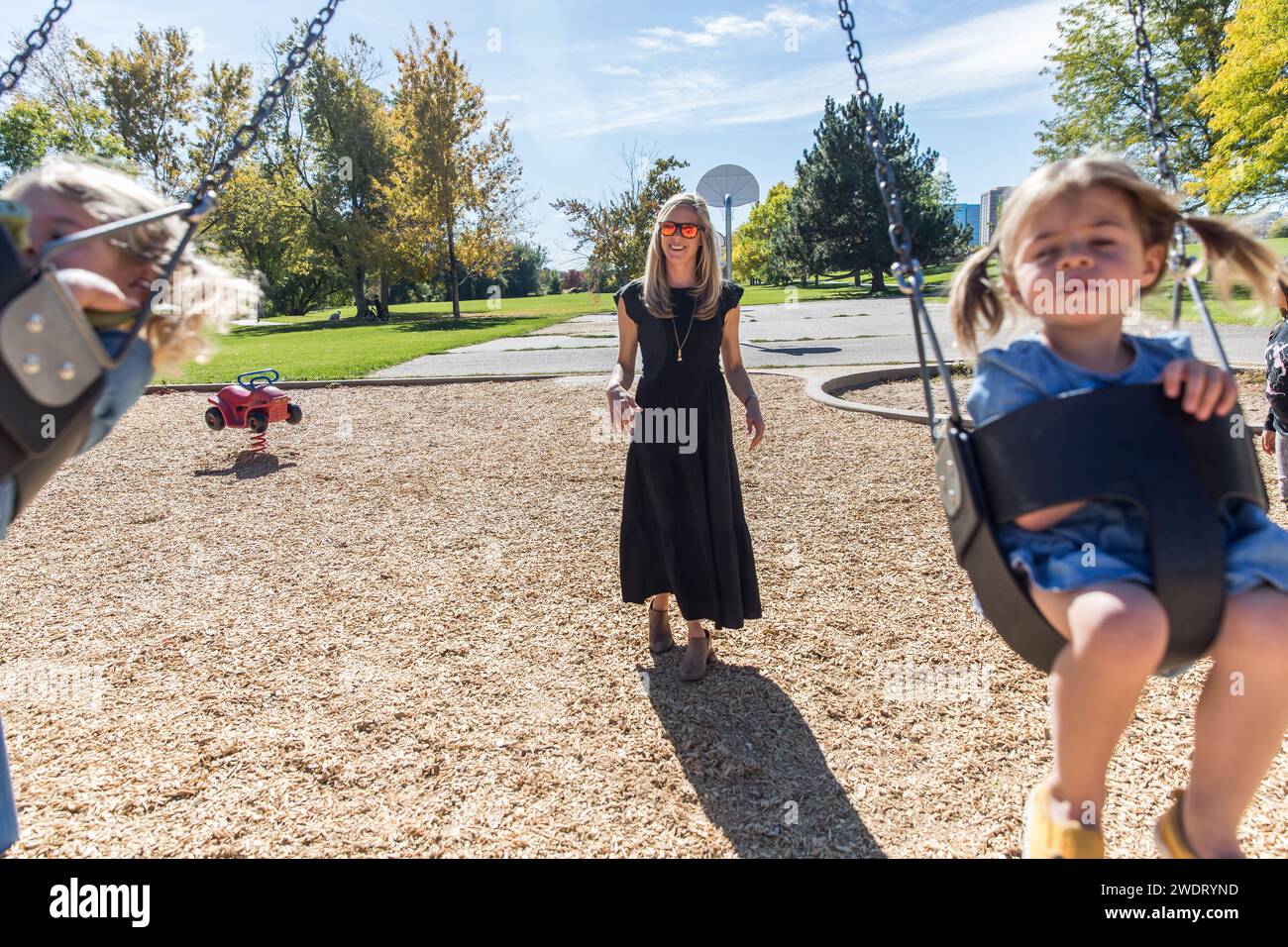 Mamma bionda che spinge le figlie sugli altalene al parco giochi Foto Stock