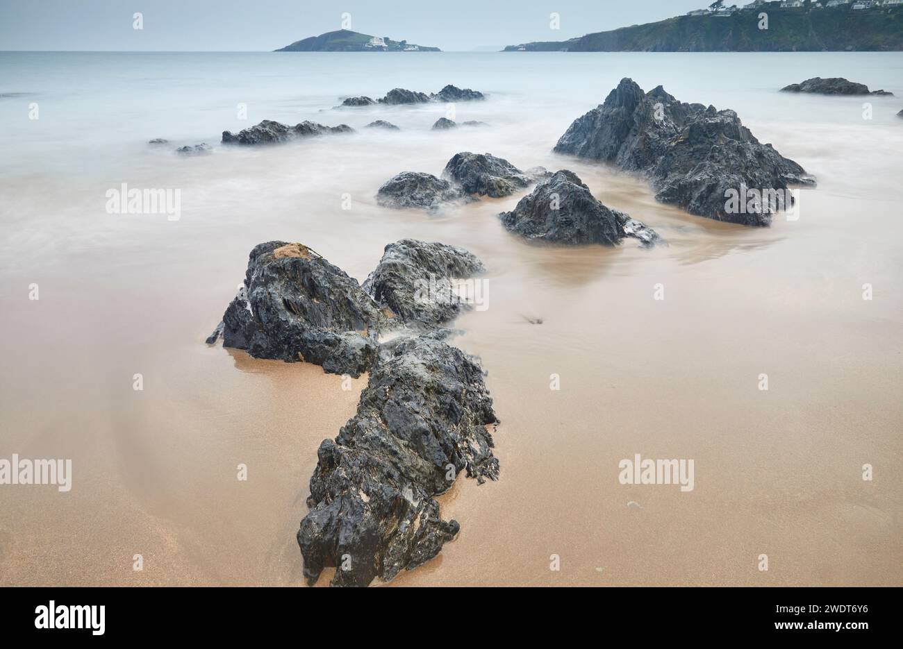 I massi della costa sepolti nella sabbia affrontano un'avanzata marea al crepuscolo, guardando dalla spiaggia di Bantham verso Burgh Island, sulla costa meridionale del Devon Foto Stock