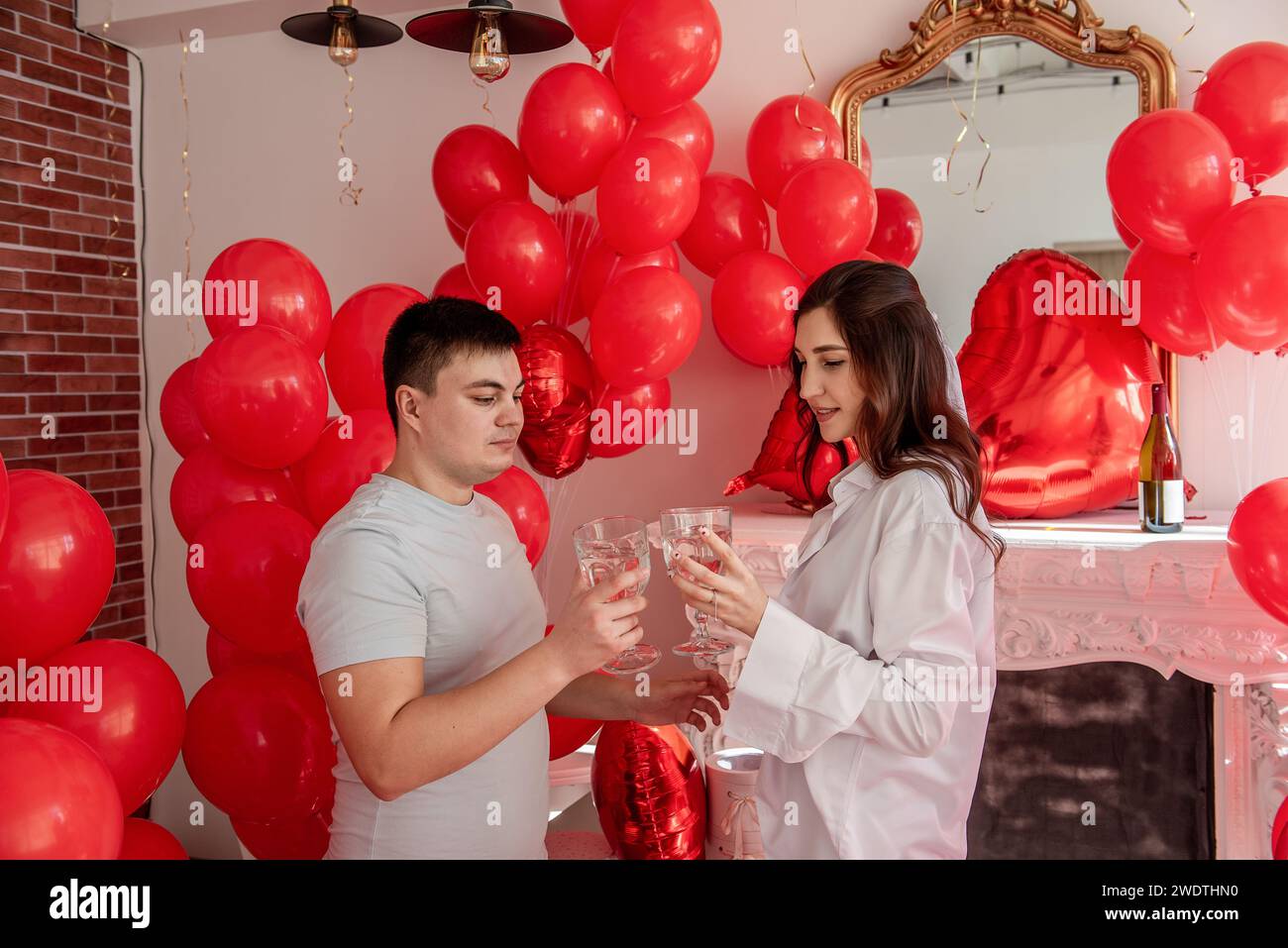 Un momento di gioia tra giovani coppie che festeggiano con un brindisi di San Valentino vicino a palloncini rossi, caminetto bianco. Donna che ride, tiene il bicchiere, sta per C. Foto Stock