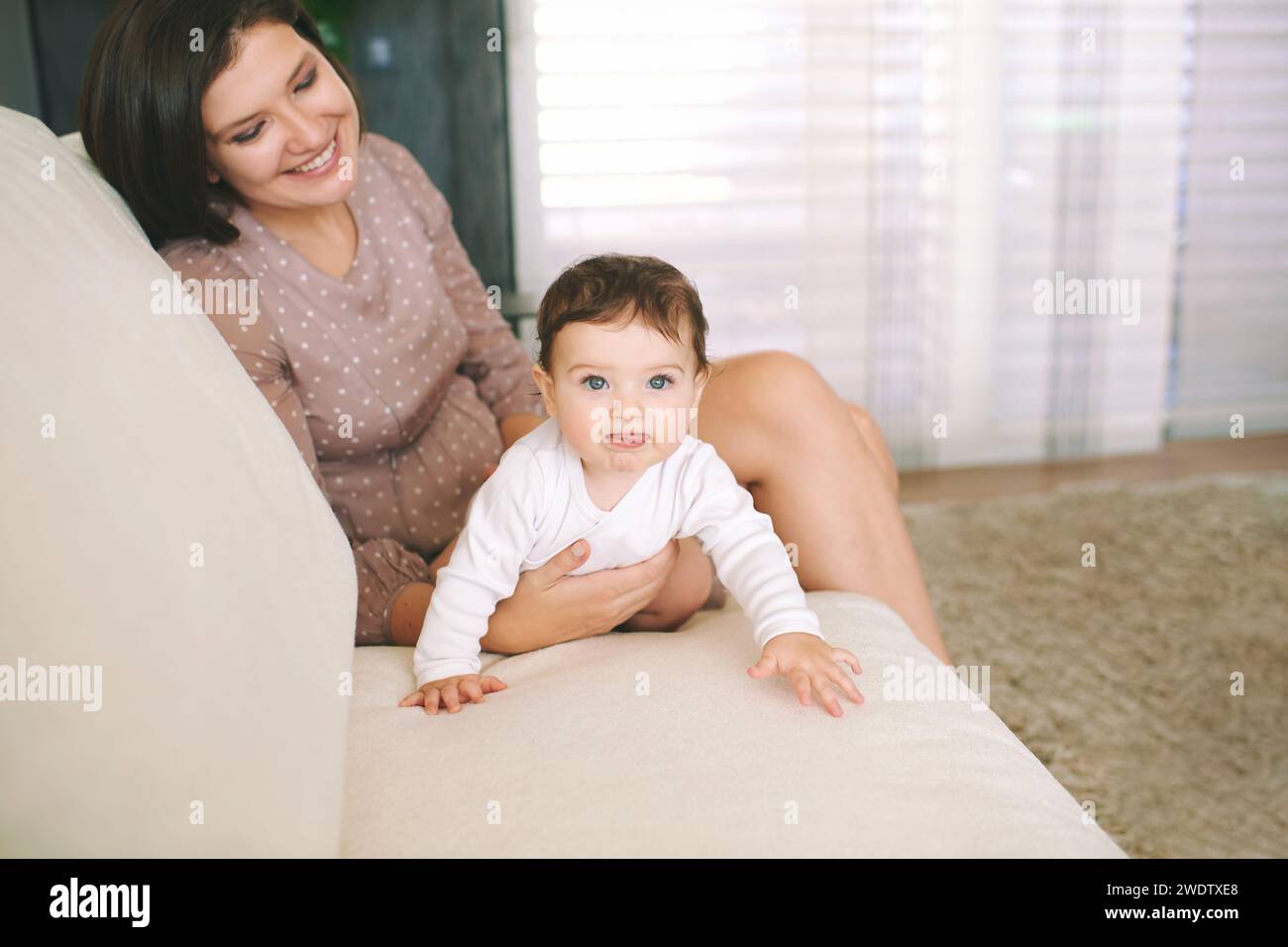 Ritratto al coperto di una giovane madre felice che gioca con un adorabile bambino, coccolandosi su un comodo divano bianco in una stanza amorevole Foto Stock