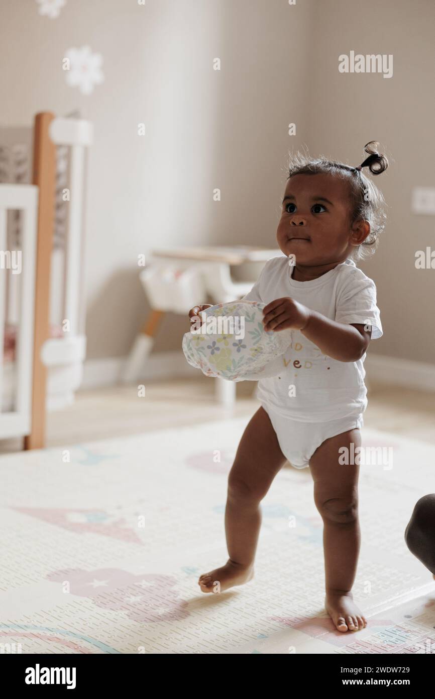 Bambina con pannolino in mano nella cameretta. Bambina afro-caucasica. Concetto di famiglia interrazziale. Foto Stock