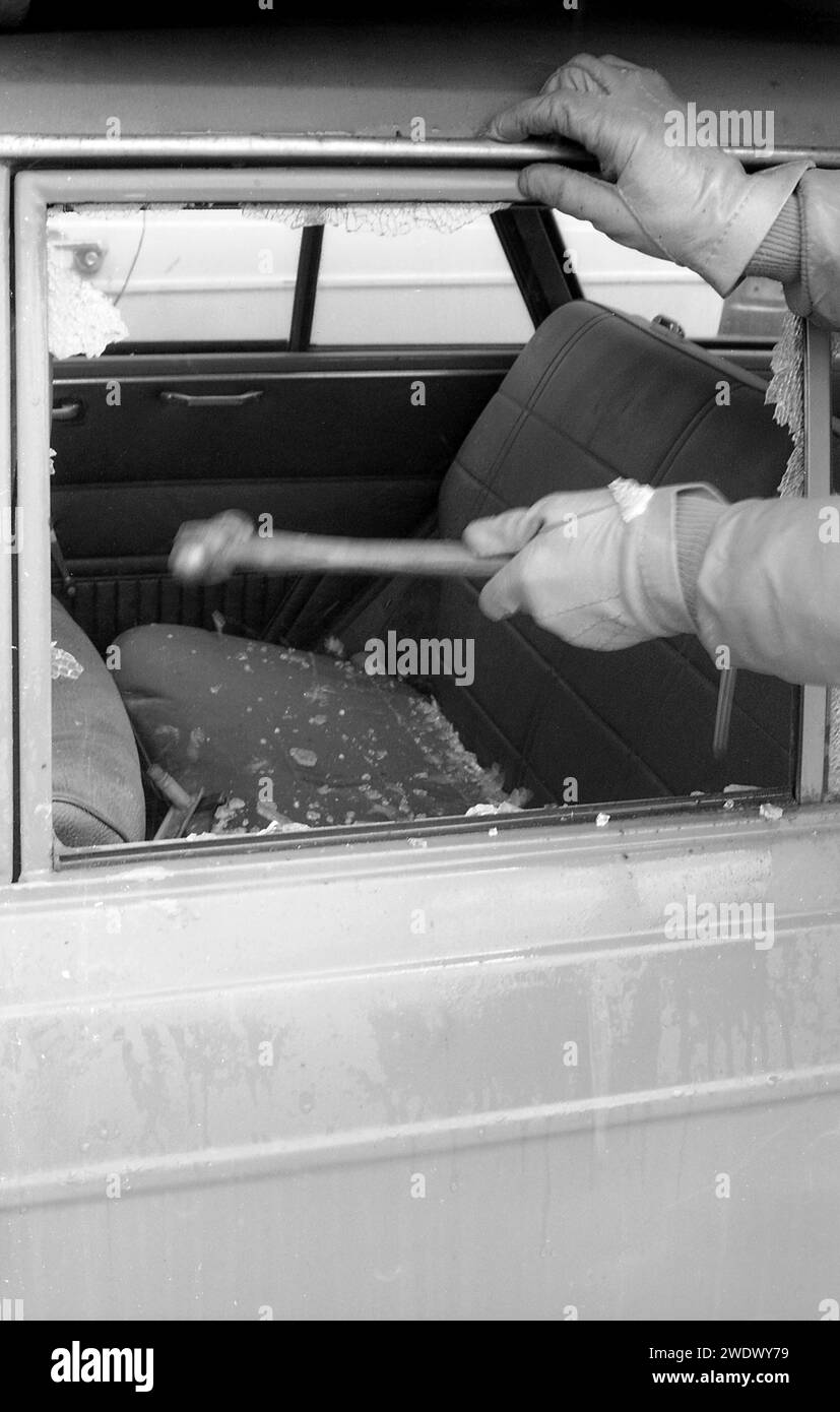 La foto storica degli anni '1970 mostra un finestrino rotto, con vetri frantumati sui sedili posteriori e le mani con guanti di un ladro d'auto che tiene un martello, Inghilterra, Regno Unito. Foto Stock
