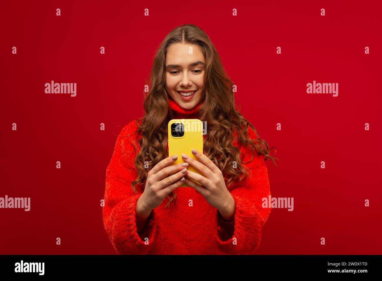 Ritratto di una giovane donna emotiva sorridente con capelli ricci Generation Z in un maglione rosso utilizzando uno smartphone moderno su uno sfondo rosso scattato in studio Foto Stock