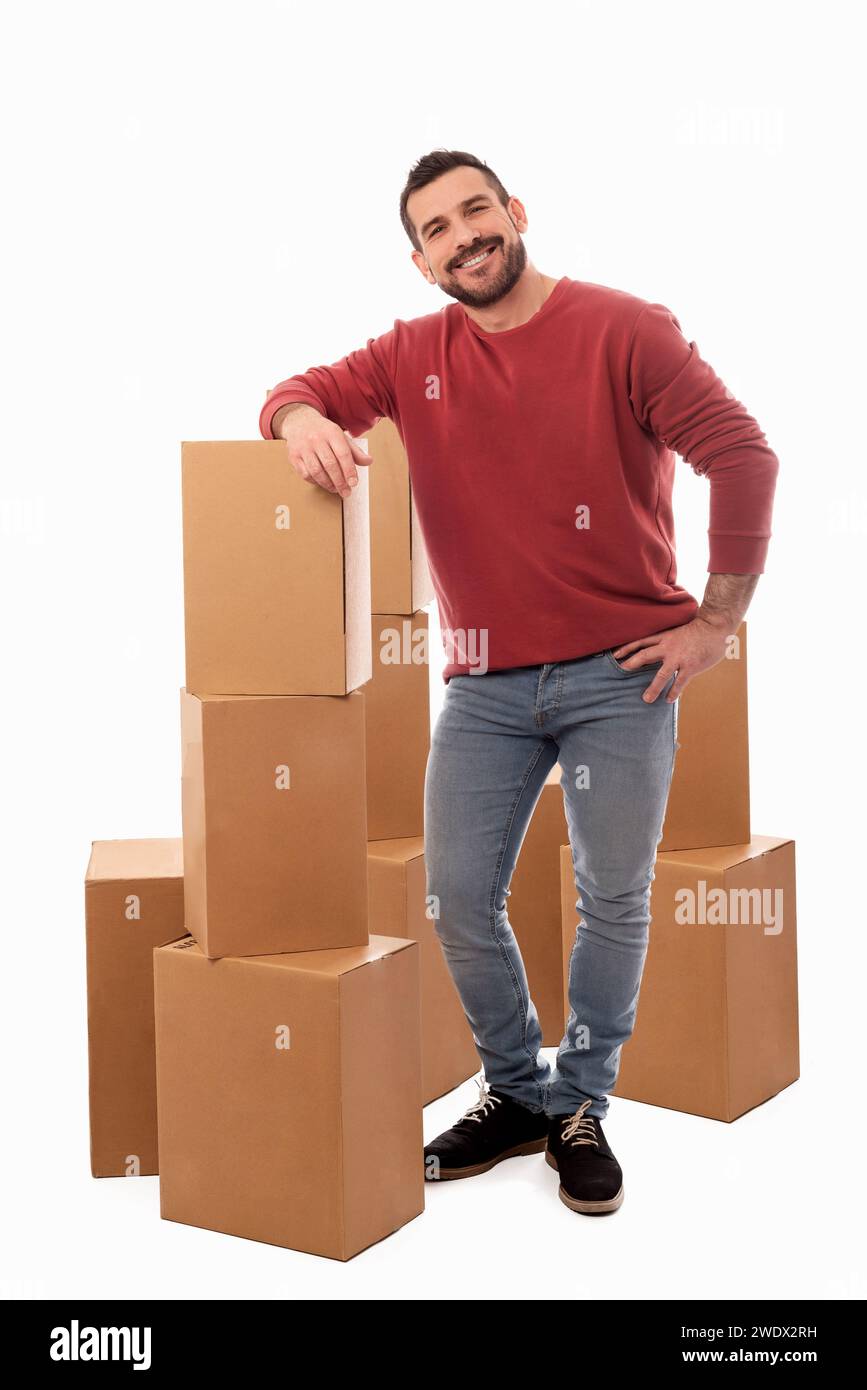Uomo sorridente in piedi con barba e maglione rosso su sfondo bianco circondato da grandi scatole di cartone. Servizio di consegna a domicilio. Spostamento del servizio di assistenza Foto Stock