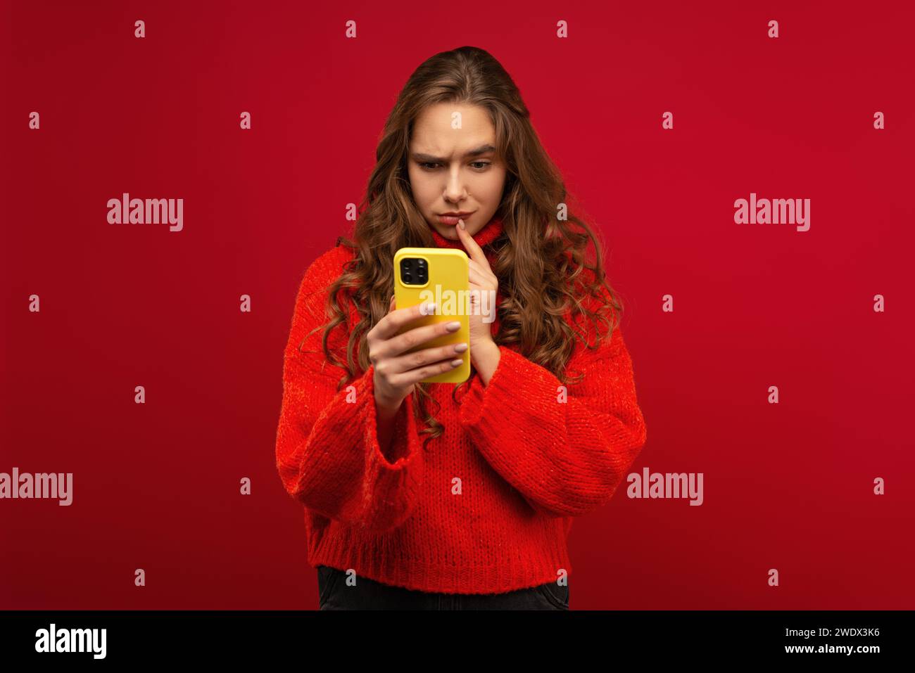 Ritratto di una giovane donna emotiva sorridente con capelli ricci Generation Z in un maglione rosso utilizzando uno smartphone moderno su uno sfondo rosso scattato in studio Foto Stock