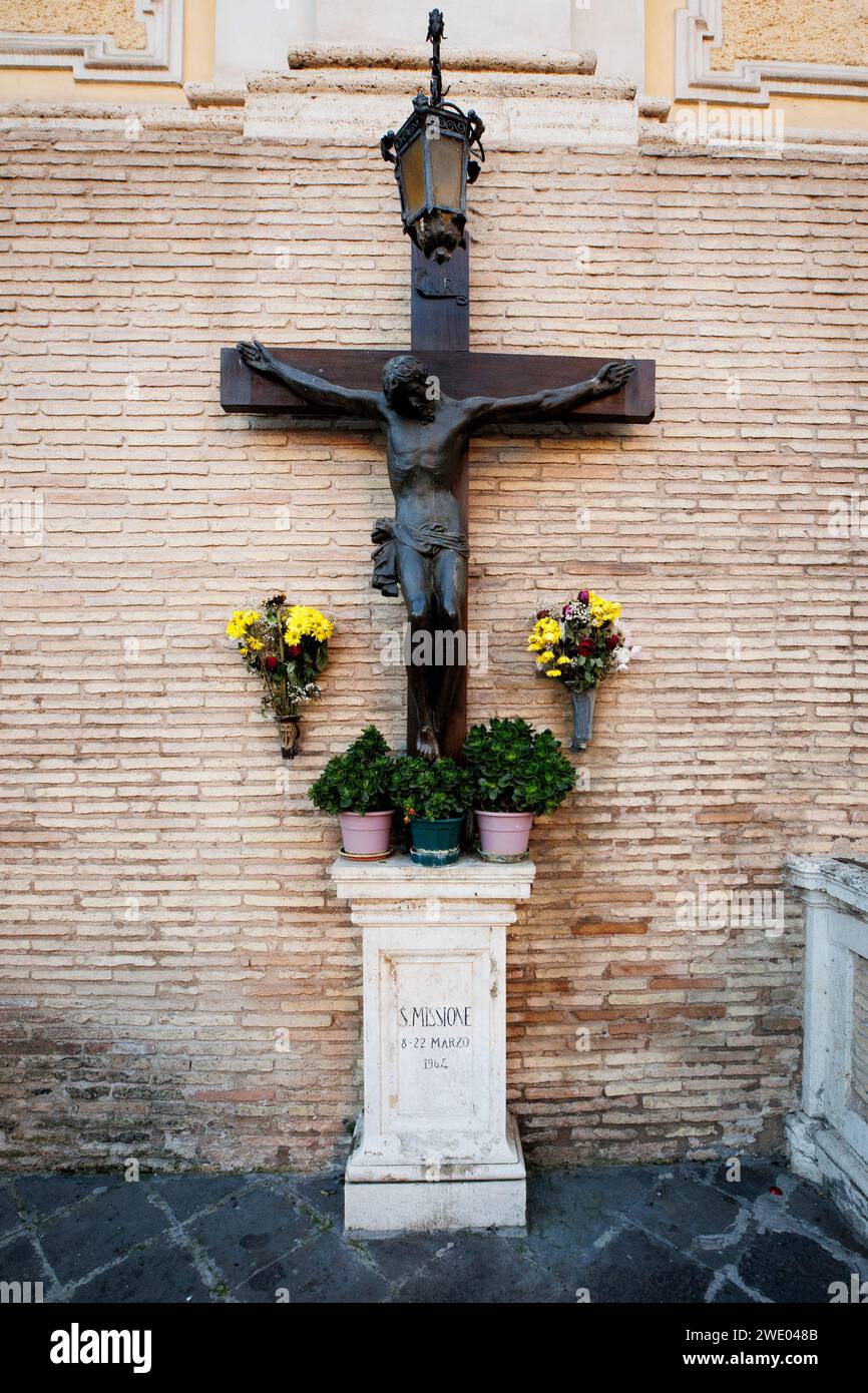 Crocifisso in bronzo con tributo floreale su una cinta muraria storica a Santa Maria delle Fornaci, Roma, simbolo della fede e della memoria Foto Stock