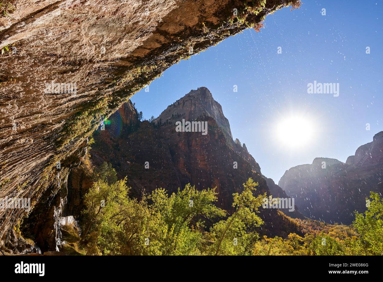 Gocce d'acqua da una sporgenza di roccia. Il sole basso brilla sul canyon, retroilluminando gli alberi autunnali e le goccioline d'acqua. La valle è circondata da roccia pe Foto Stock