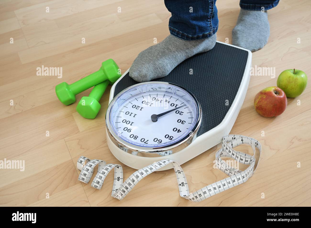 Piedi di un uomo in calzini grigi che calpestano una bilancia analogica, manubri verdi, metro a nastro e mele che giacciono nelle vicinanze su un pavimento di legno, fitness e.. Foto Stock