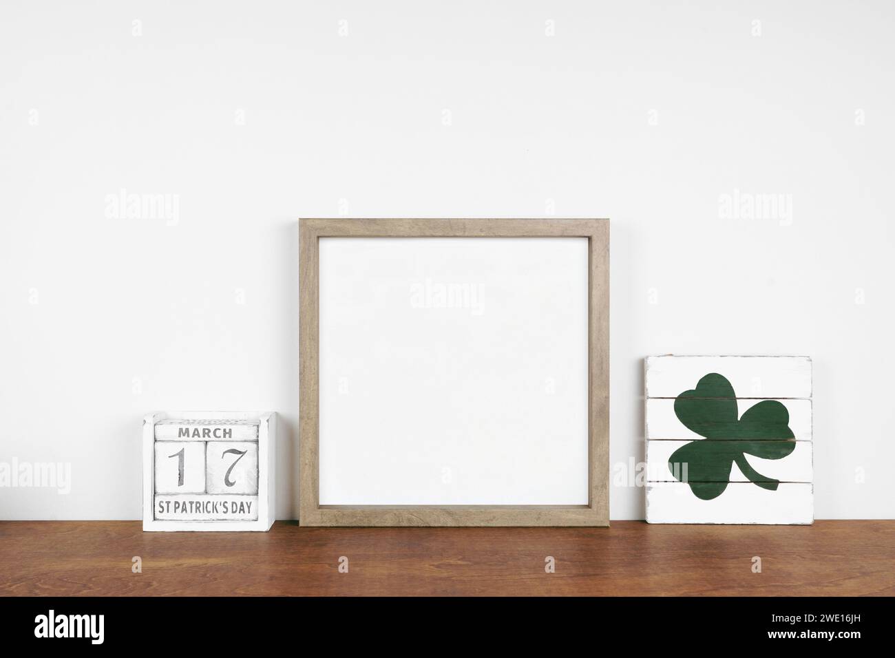 Montatura in legno con decorazioni St Patricks Day su un ripiano in legno. Cartellone e calendario in legno squallido ed elegante. Cornice quadrata contro una parete bianca. Copia spazio. Foto Stock