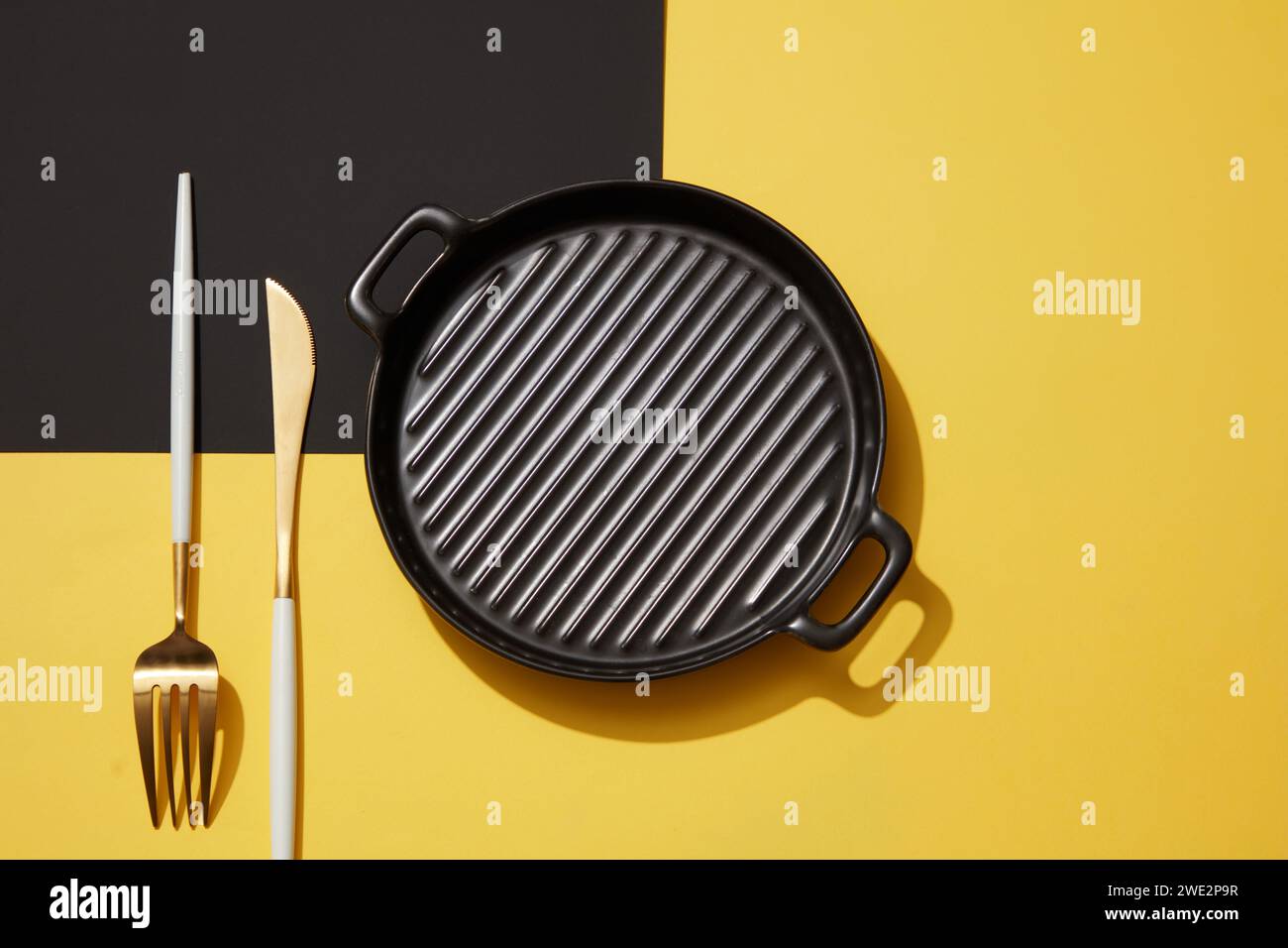 Un recipiente nero vuoto posizionato su sfondo nero e giallo con un set di coltello e forchetta. Vista dall'alto. Regole di etichetta. Scena minima astratta per un Foto Stock