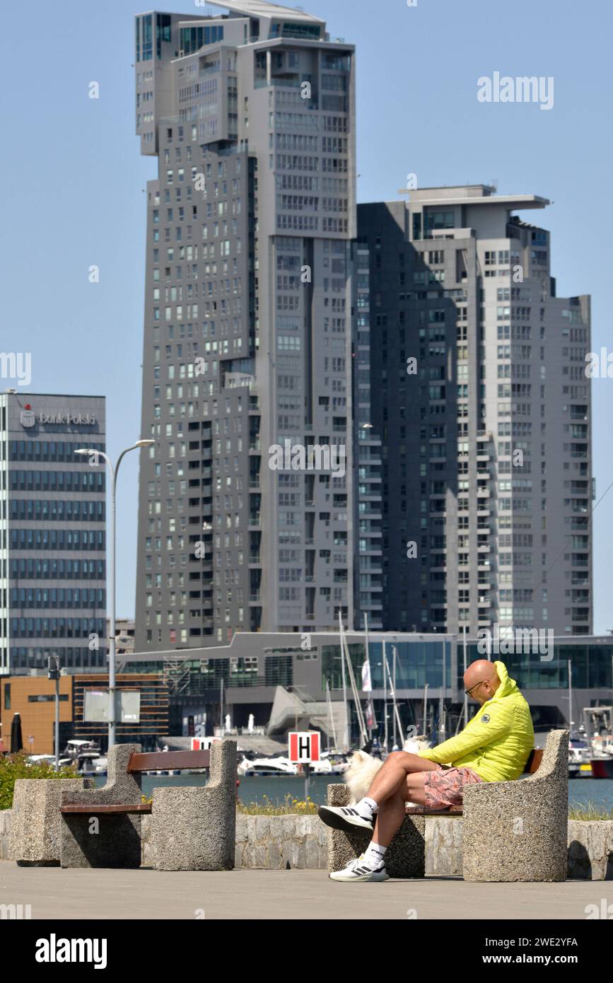 Uomo del posto che indossa una giacca gialla e un piccolo cane seduto su una panchina presso la Marina e il porto di Gdynia sul Mar Baltico a Gdynia, Polonia, Europa, UE Foto Stock