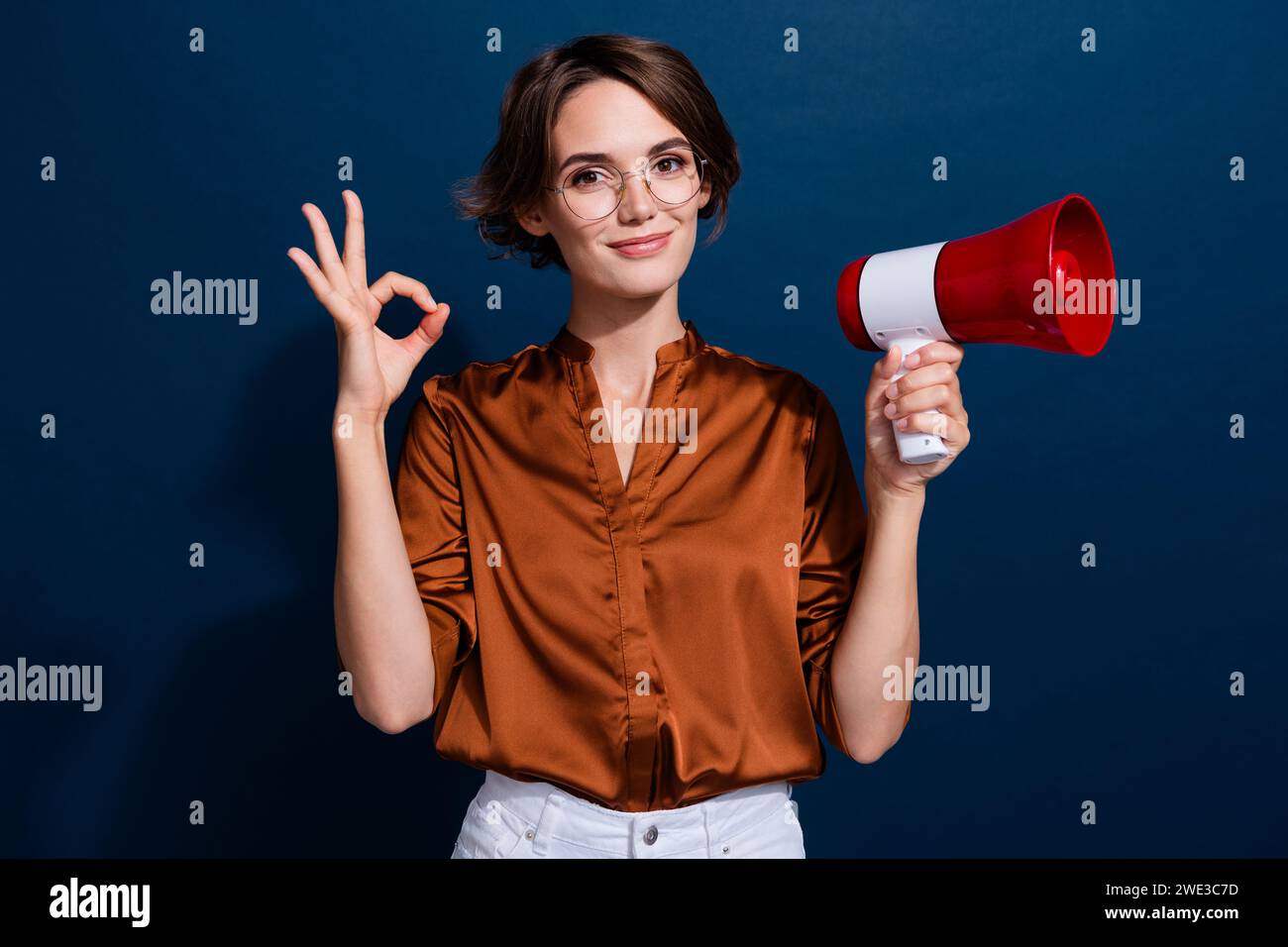 Ritratto fotografico di una giovane donna attraente che mostra un altoparlante con il simbolo dell'occhiello vestito da elegante blusa marrone isolata su sfondo marrone scuro Foto Stock