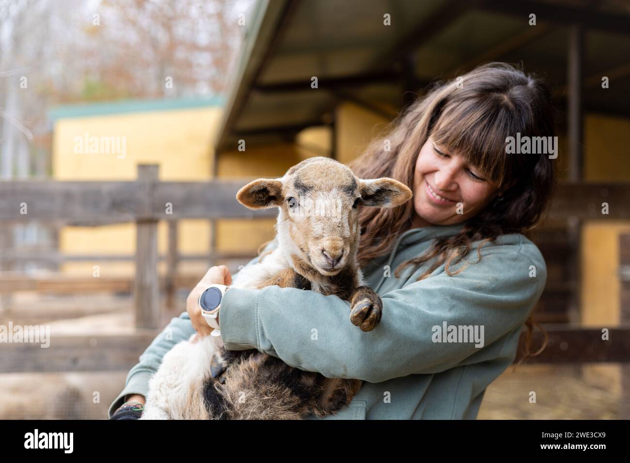 Felice giovane contadina che abbraccia una piccola pecora in una fattoria rurale biologica. Benessere e cura degli animali in un fienile. Foto Stock