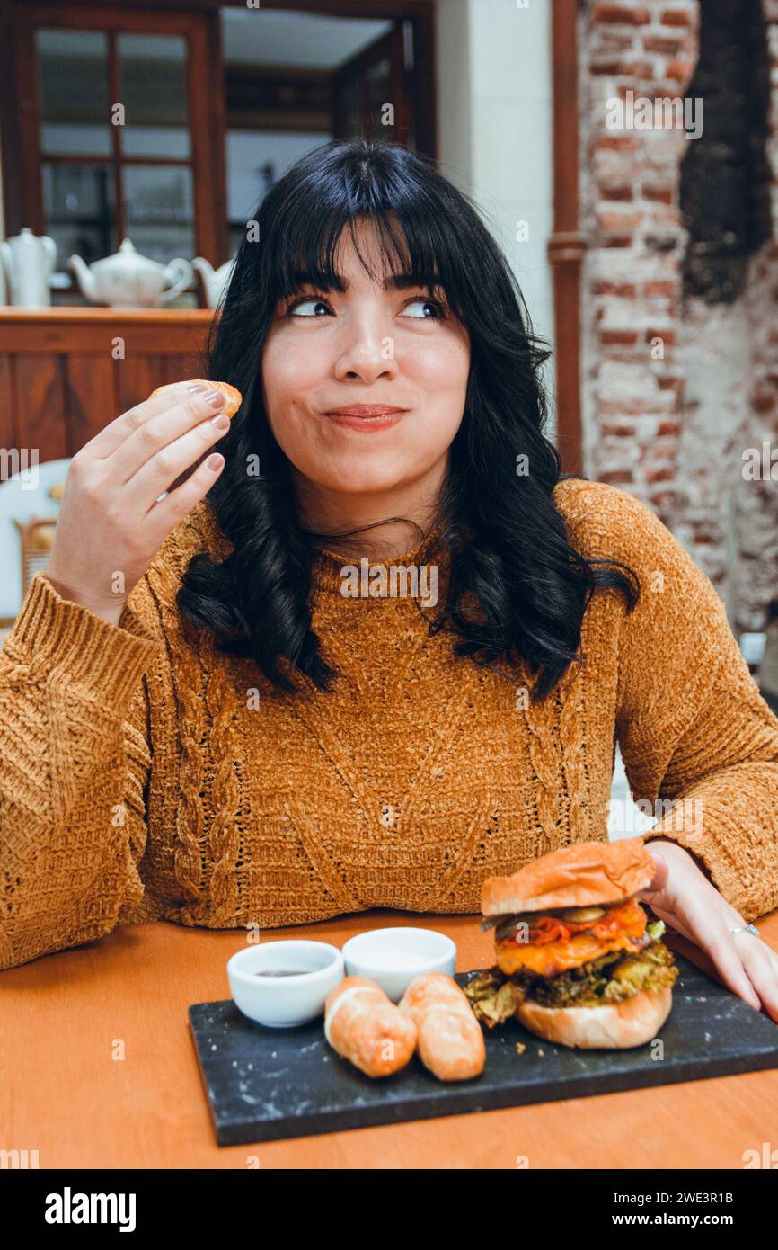 Immagine verticale di una giovane latina con capelli neri e abiti marroni, happy sitting nel ristorante che mangia tequenos, cibo tradizionale venezuelano, con hambu Foto Stock
