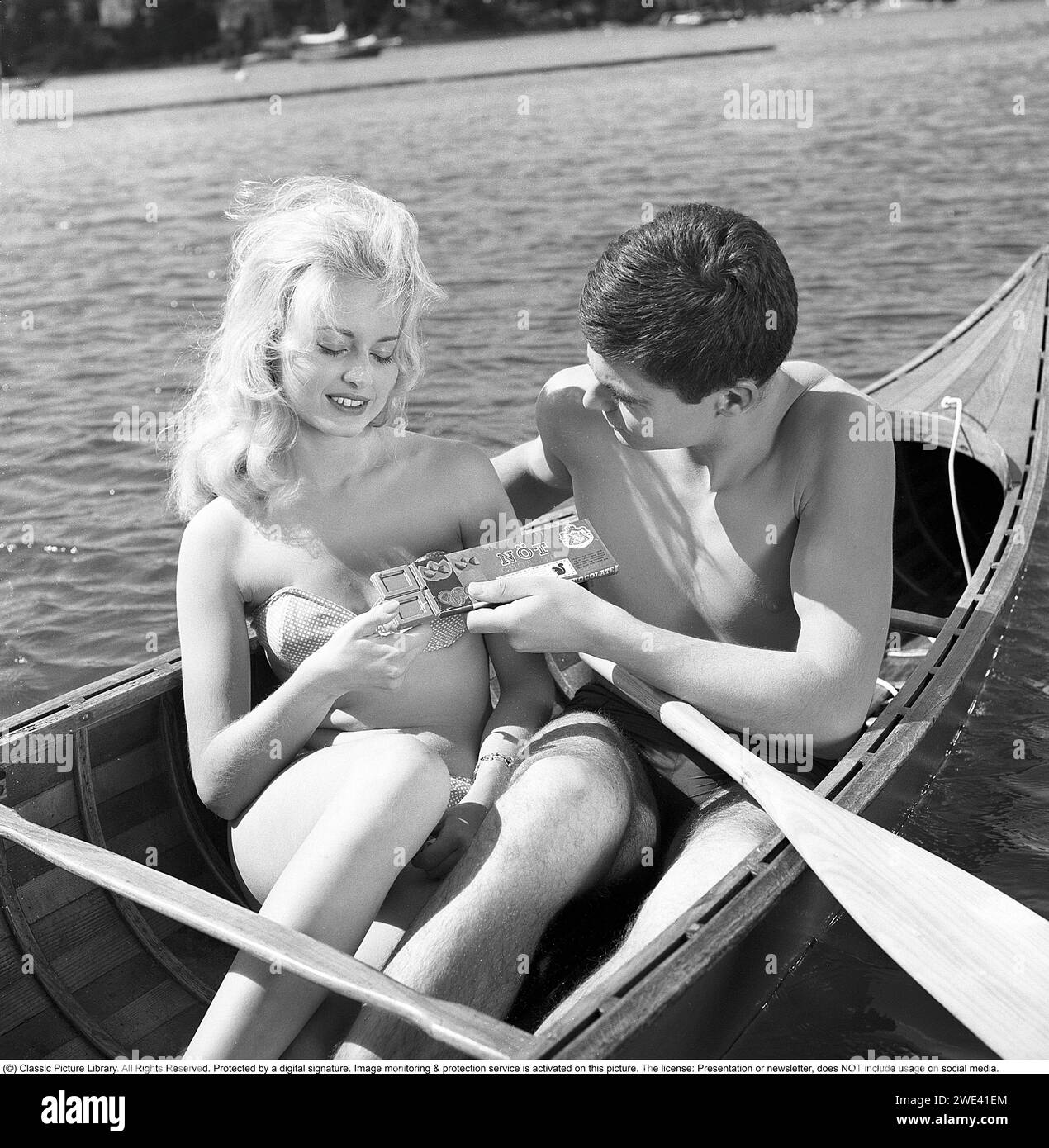 Mazetti Chocolate. Una giovane coppia siede insieme in canoa in una soleggiata giornata estiva e lui le offre il cioccolato dalla sua barretta di cioccolato. Svezia 1962. Kristoffersson rif. CZ54 Foto Stock