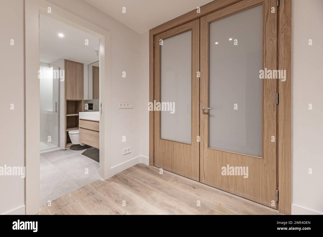 Un piccolo bagno dal design moderno con mobili sospesi in legno con accesso da un distributore con porte in legno di quercia e vetro Foto Stock