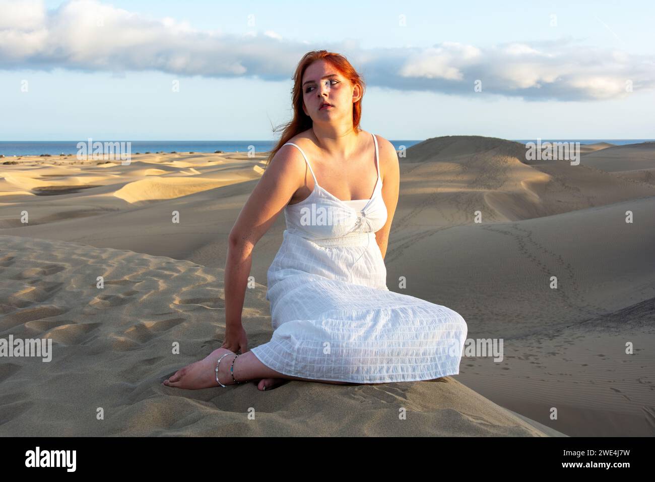 Una giovane bella ragazza dai capelli rossi con un vestito bianco siede nelle dune di sabbia nel tardo pomeriggio e gode degli ultimi raggi di sole Foto Stock
