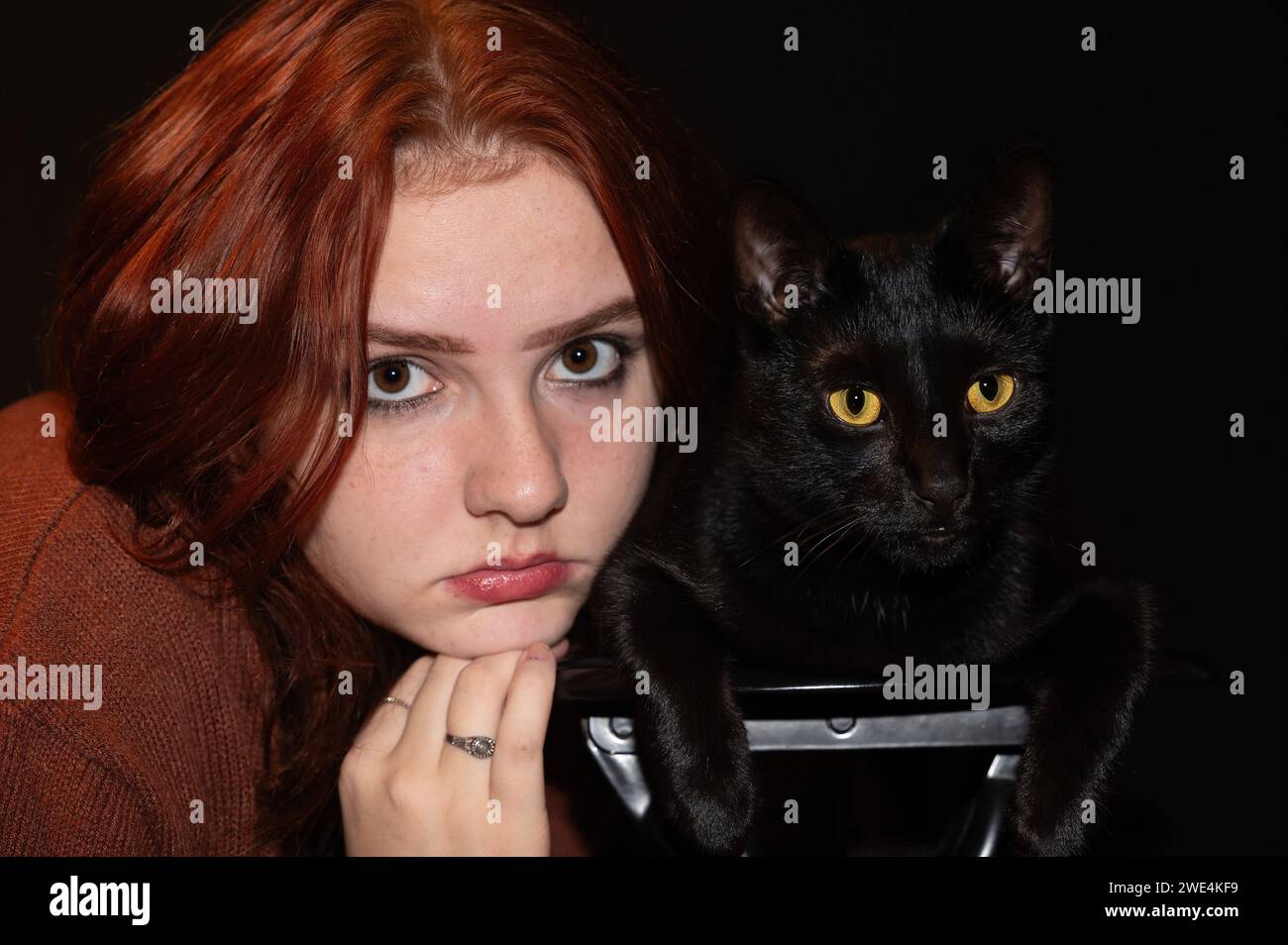 Una ragazza dai capelli rossi con il suo gatto nero e sfondo nero, entrambi guardano avanti Foto Stock
