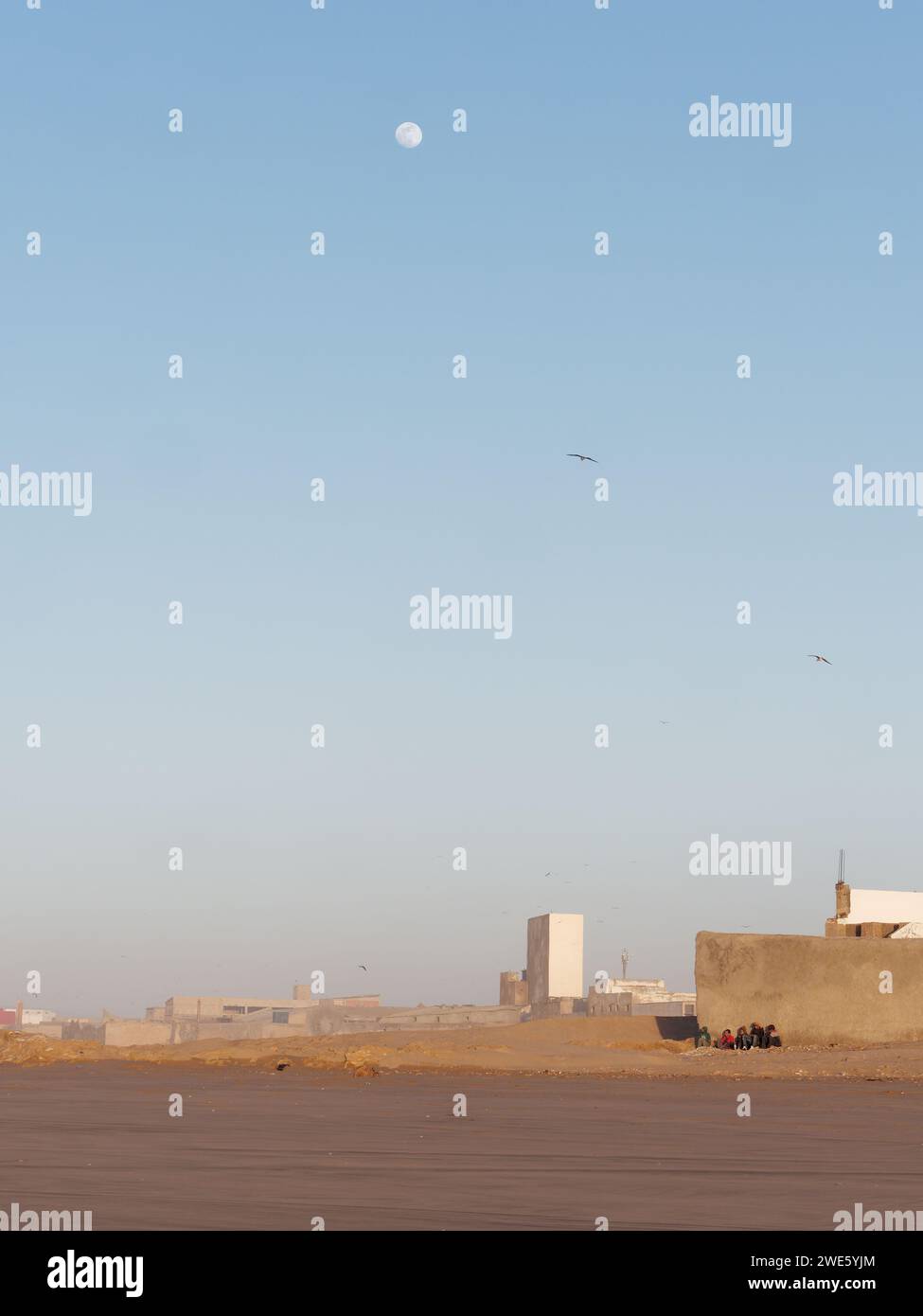 Un gruppo di uomini siede sul bordo di una spiaggia sabbiosa con la luna sopra nella parte industriale di Essaouira con edifici insoliti, Marocco, 23 gennaio 2024 Foto Stock