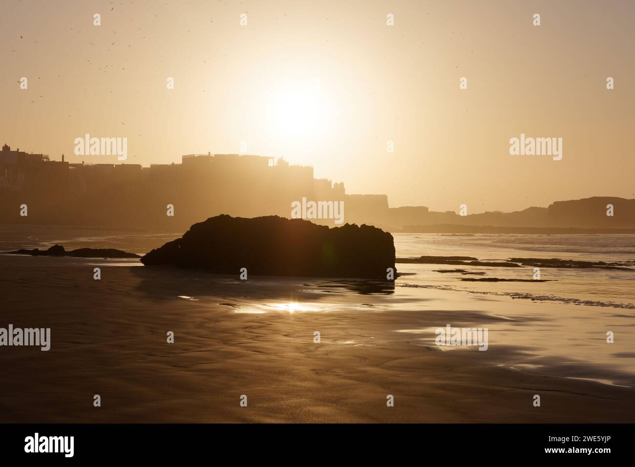Affioramento roccioso su una spiaggia sabbiosa al tramonto con il sole sulla medina di Essaouira, Marocco, 23 gennaio 2024 Foto Stock