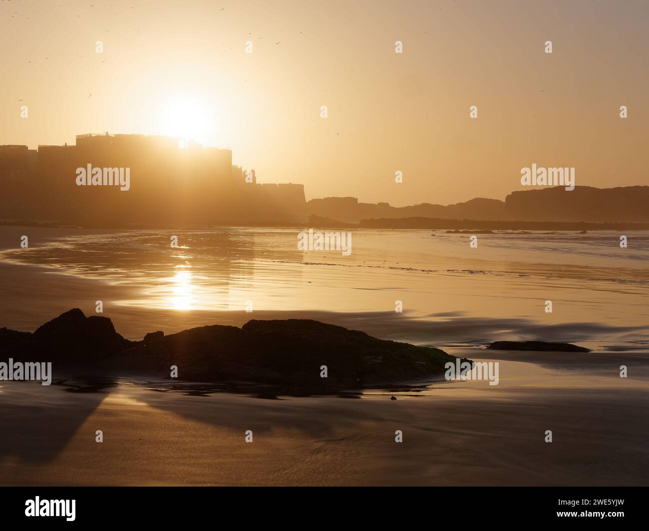 Affioramento roccioso su una spiaggia sabbiosa al tramonto con il sole sulla medina di Essaouira, Marocco, 23 gennaio 2024 Foto Stock