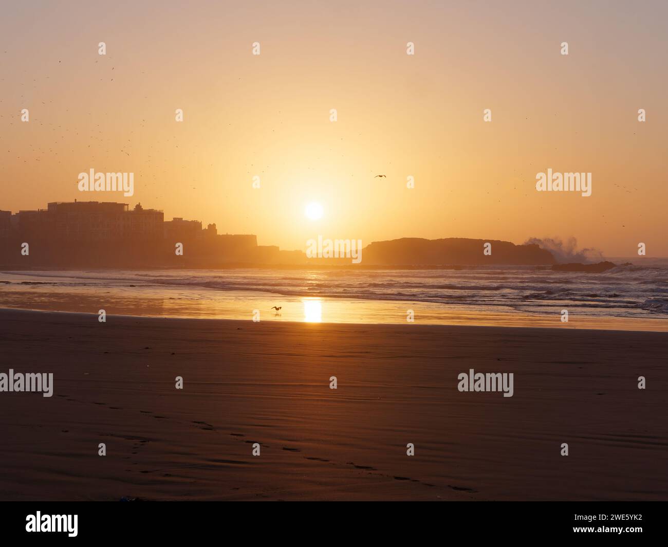 Le onde si infrangono su una roccia al tramonto su una spiaggia sabbiosa con medina a sinistra, a Essaouira, Marocco, 23 gennaio 2024 Foto Stock