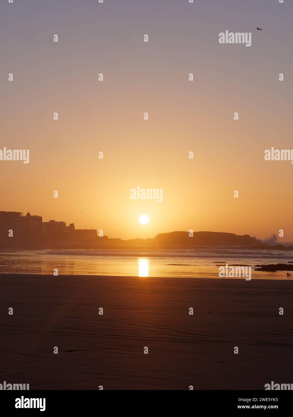 Le onde si infrangono su una roccia al tramonto su una spiaggia sabbiosa con medina a sinistra, a Essaouira, Marocco, 23 gennaio 2024 Foto Stock