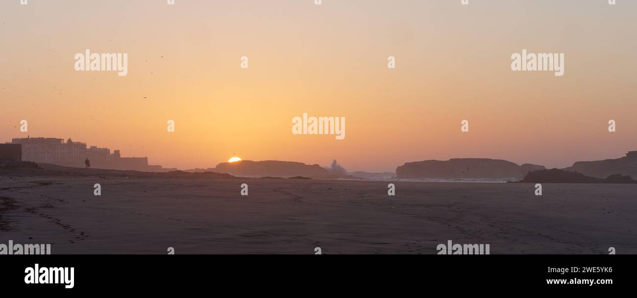 La figura solitaria cammina su una spiaggia sabbiosa al tramonto mentre le onde si schiantano contro una roccia con la Medina a sinistra, Essaouira, Marocco, 23 gennaio 2024 Foto Stock