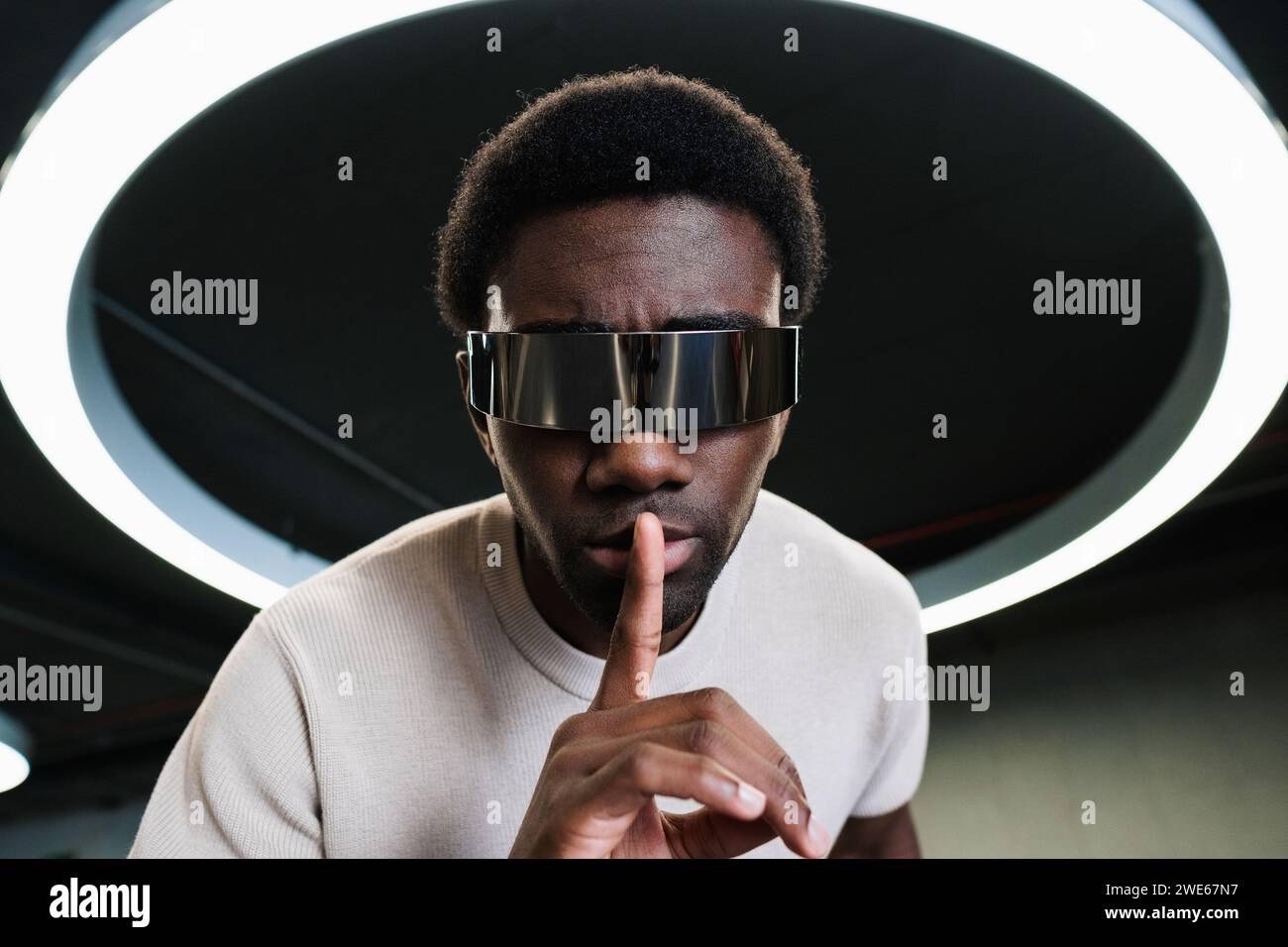 Giovane con futuristici bicchieri cyber sotto una moderna lampada ad anello che mette l'indice sulle labbra Foto Stock