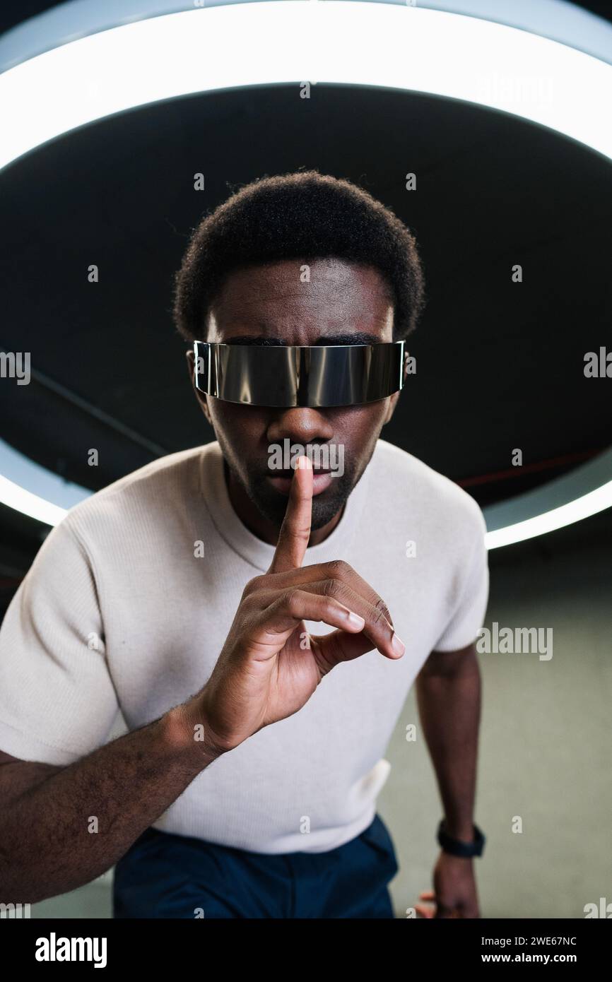 Giovane con futuristici bicchieri cyber sotto una moderna lampada ad anello che mette l'indice sulle labbra Foto Stock