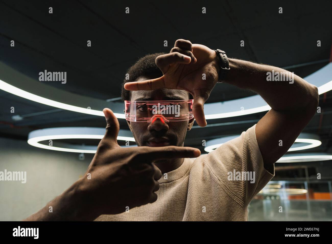 Giovane uomo con futuristici occhiali cyber sotto una moderna lampada ad anello che guarda attraverso la cornice delle dita Foto Stock