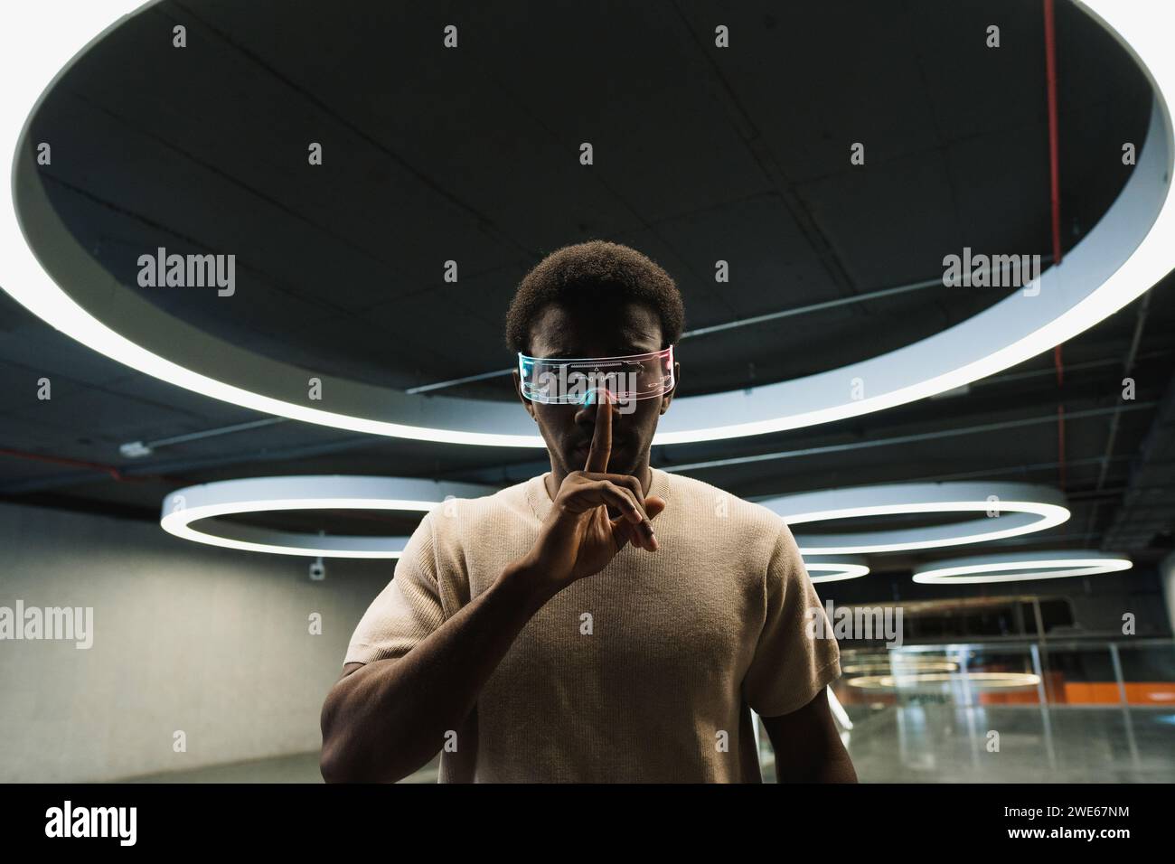 Giovane con futuristici bicchieri cyber sotto una moderna lampada ad anello che mette l'indice sulle labbra Foto Stock