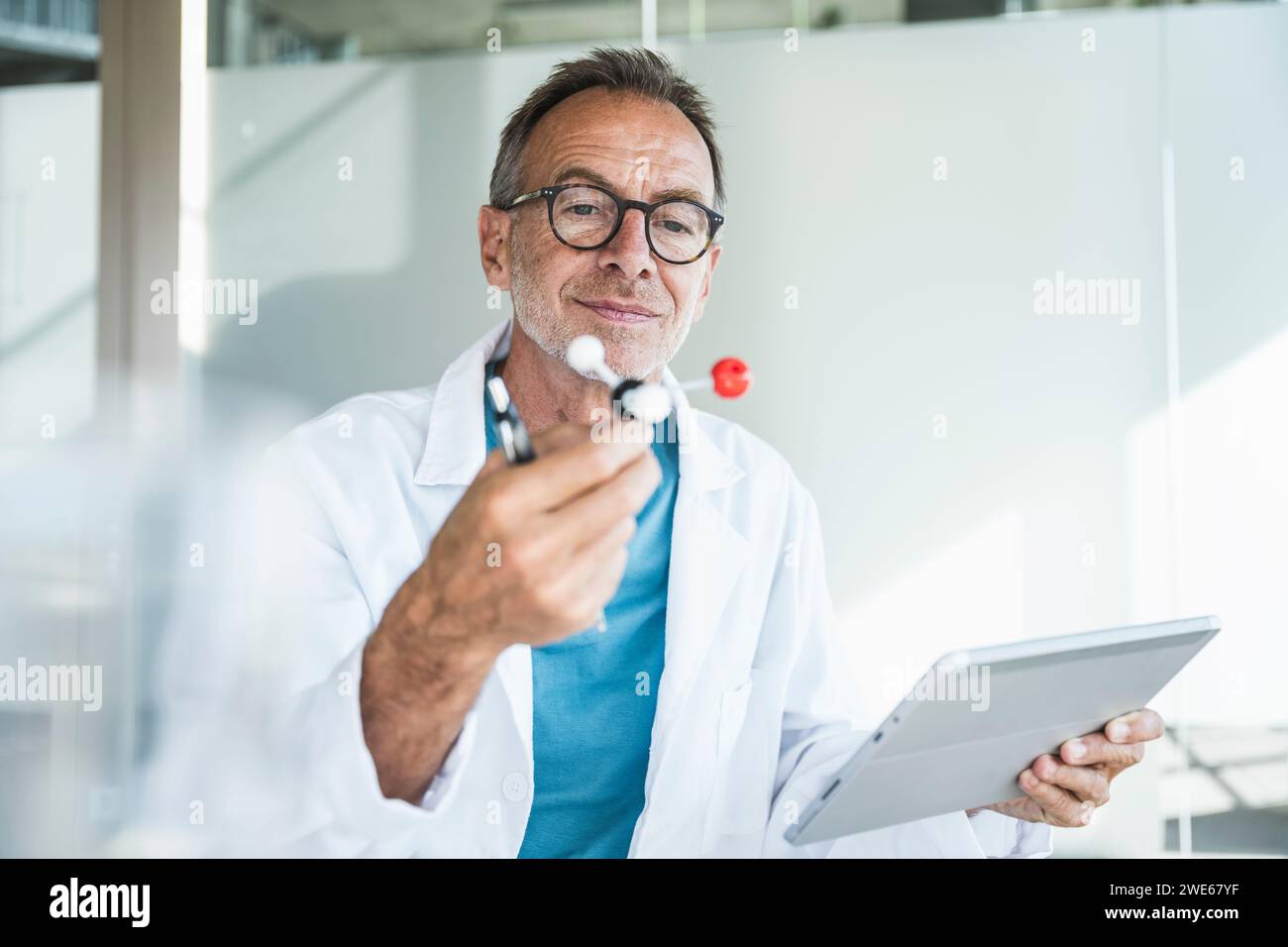 Uomo sorridente in camice da laboratorio che tiene in mano un tablet PC ed esamina la struttura molecolare Foto Stock