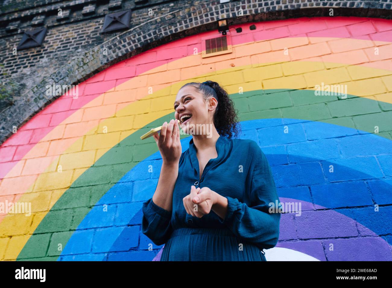 Giovane donna felice che invia messaggi vocali tramite smartphone di fronte al muro arcobaleno Foto Stock