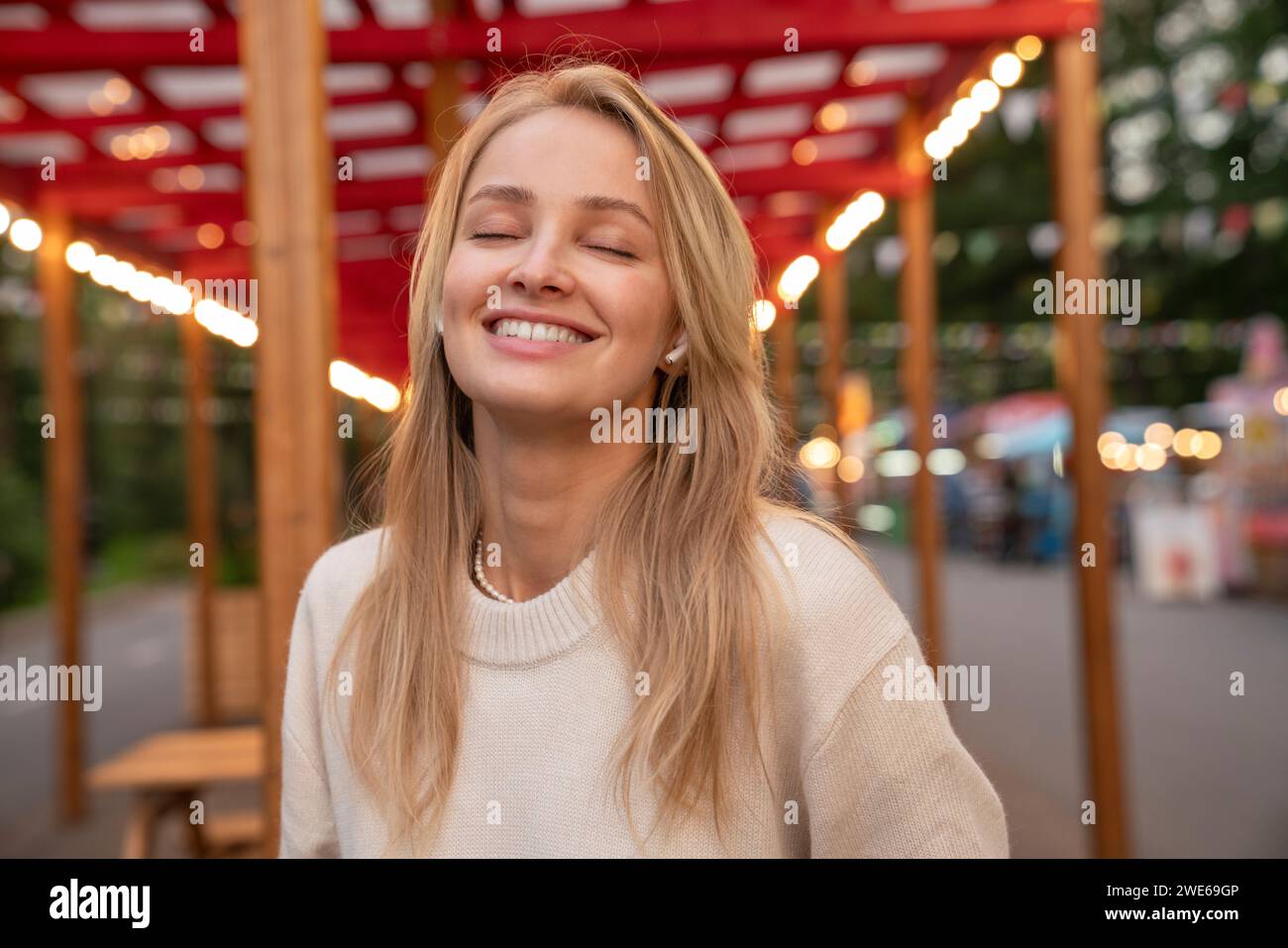 Giovane donna bionda sorridente che indossa cuffie auricolari wireless mentre ascolta musica al parco divertimenti Foto Stock