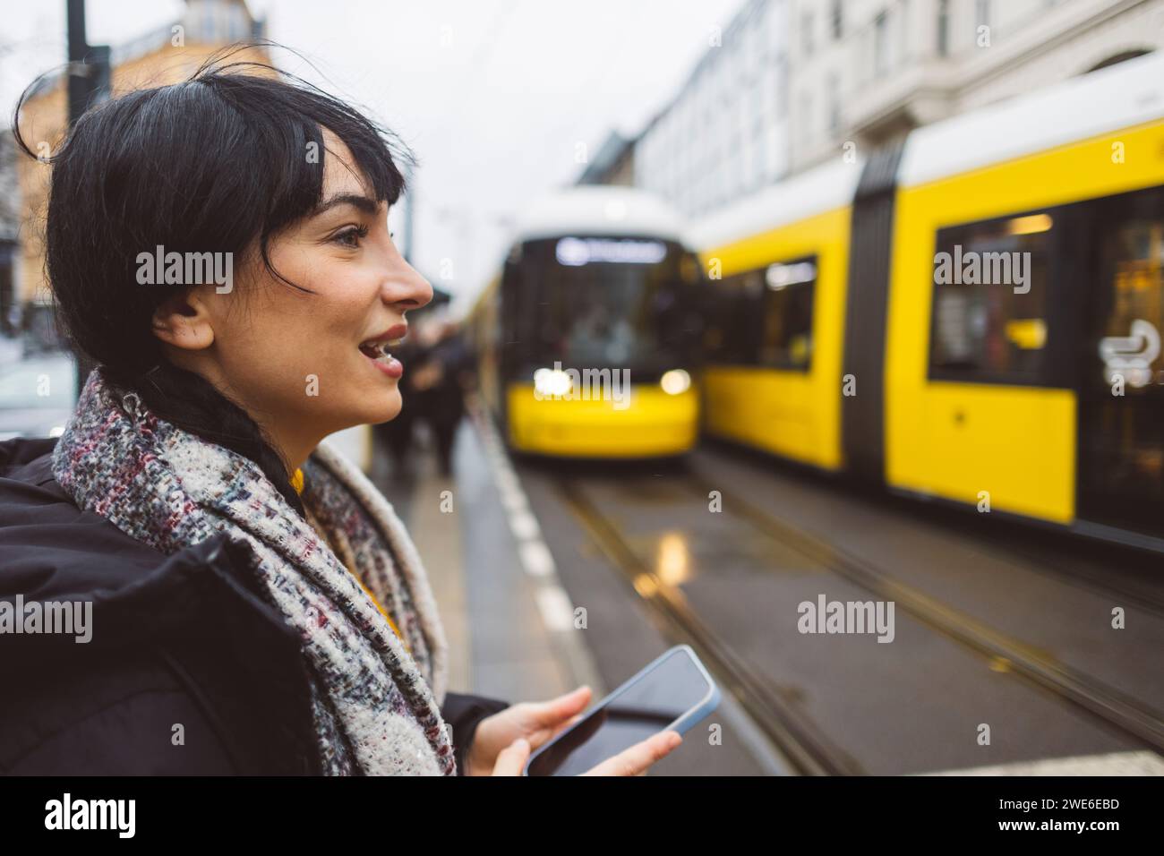 Donna felice che tiene uno smartphone e aspetta alla stazione del tram Foto Stock