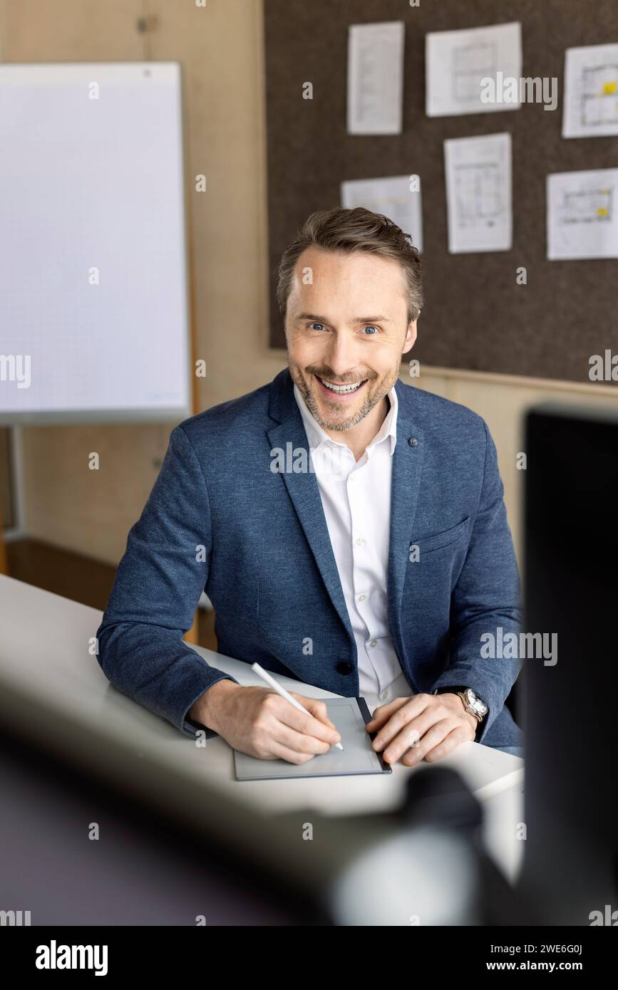 Uomo d'affari sorridente con un tablet PC seduto alla scrivania sul posto di lavoro Foto Stock