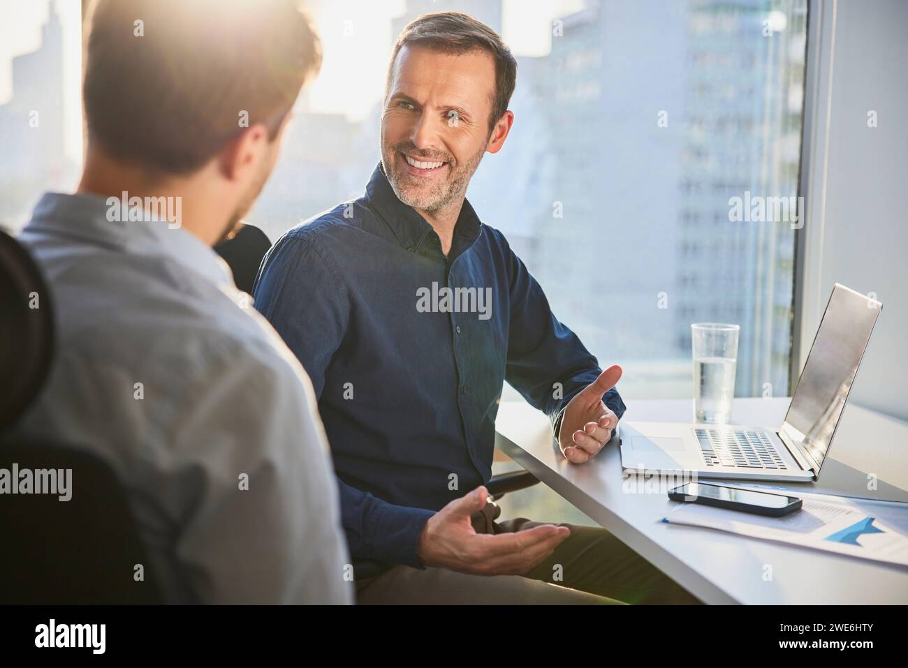 Uomo d'affari maturo e felice che condivide idee e discute con un collega alla scrivania Foto Stock