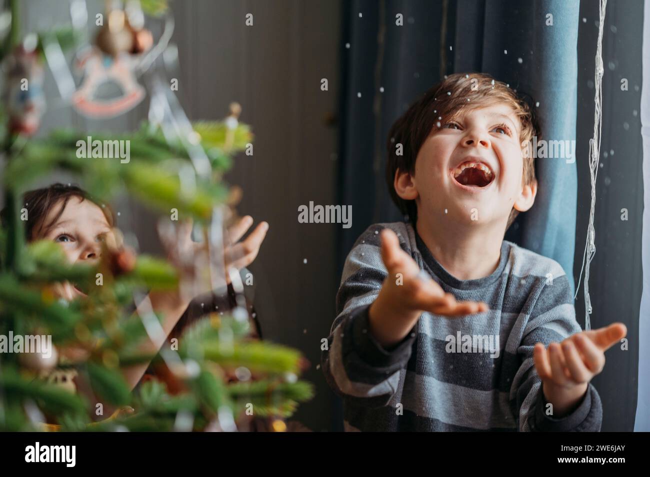 Ragazzi giocosi che prendono la neve finta vicino all'albero di Natale a casa Foto Stock