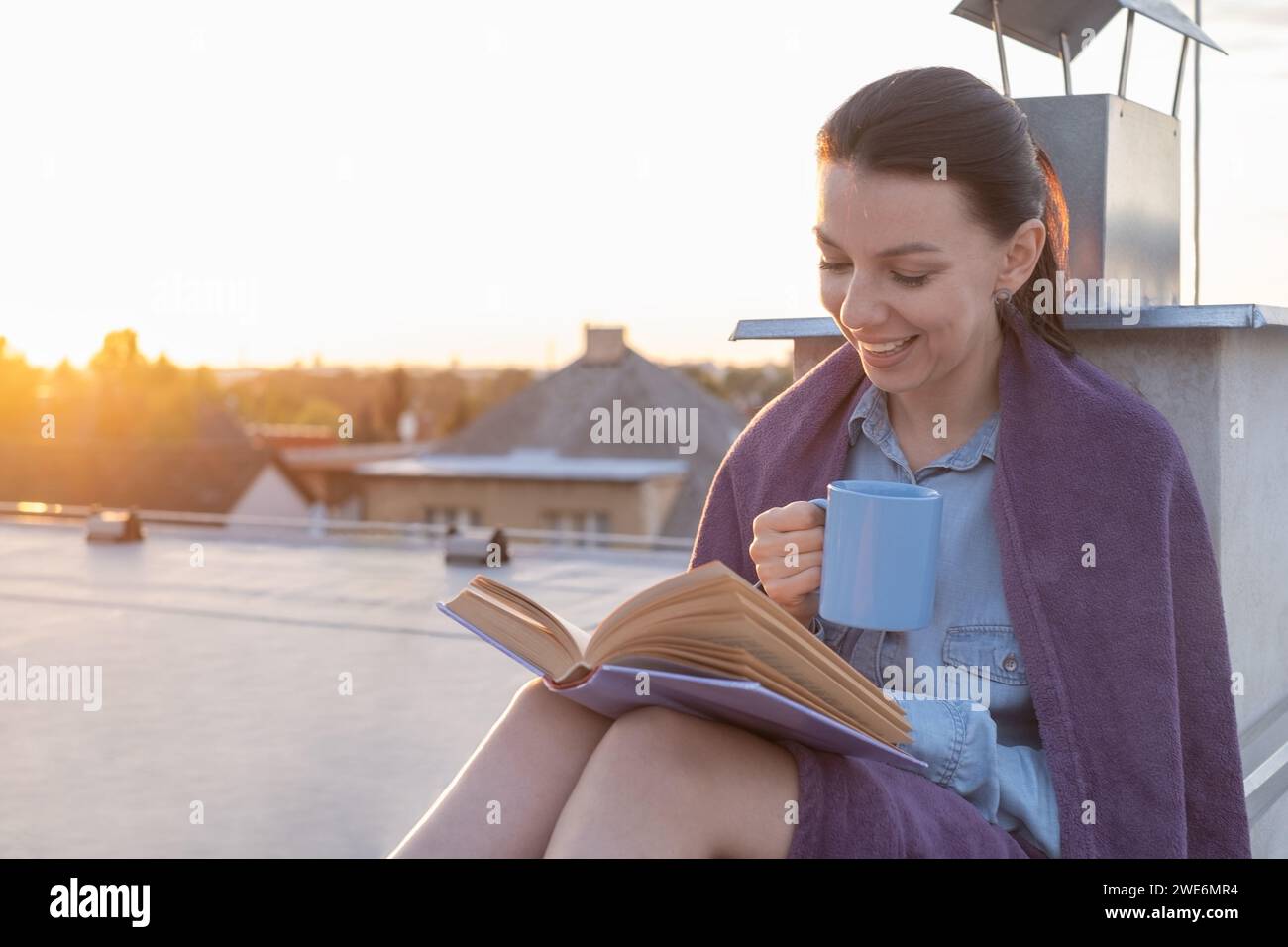 Una giovane donna ama leggere un libro con un sorriso su un tetto piatto durante l'ora d'oro. Settore delle case private, bellezza del tramonto con un caldo tramonto Foto Stock