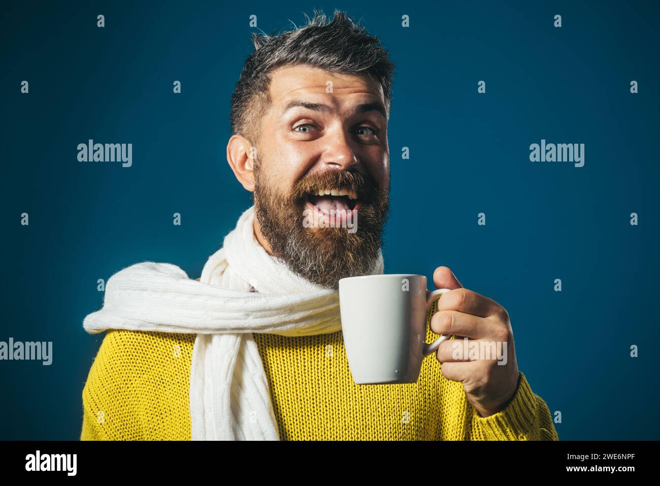 Uomo felice con la barba e l'elegante acconciatura che beve una tazza di caffè caldo o tè. Uomo barbuto in maglione giallo avvolto in sciarpa bianca per la pausa caffè Foto Stock