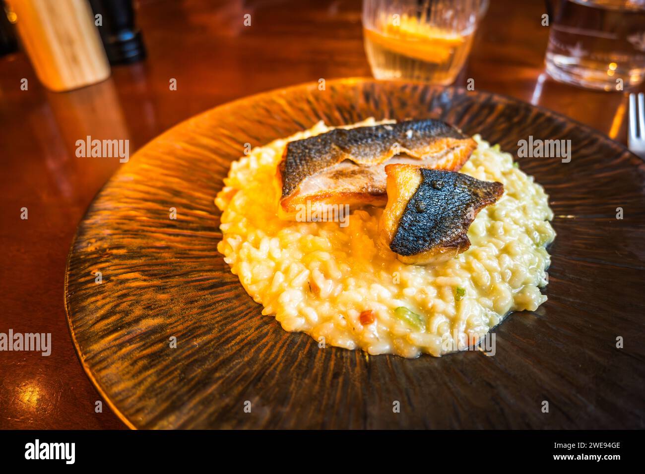 Filetto di pesce arrosto (spigola) su risotto con verdure su piatto decorativo, bevanda, bicchiere sul tavolo del ristorante. Cucina italiana. Foto Stock