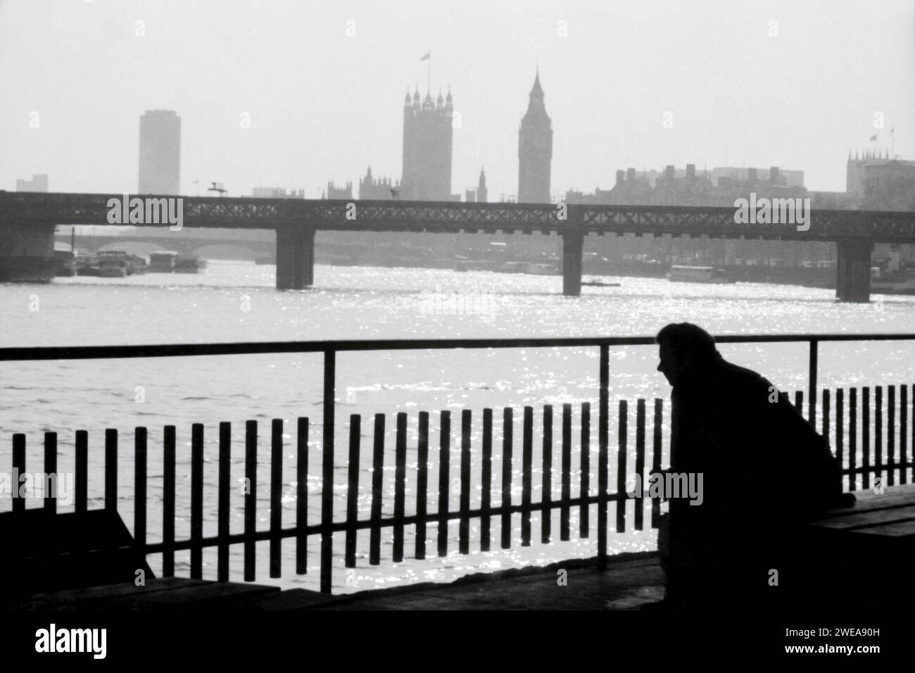 Uomo seduto accanto al Tamigi con Hungerford Bridge e Westminster sullo sfondo. Londra, circa 1981. Foto Stock