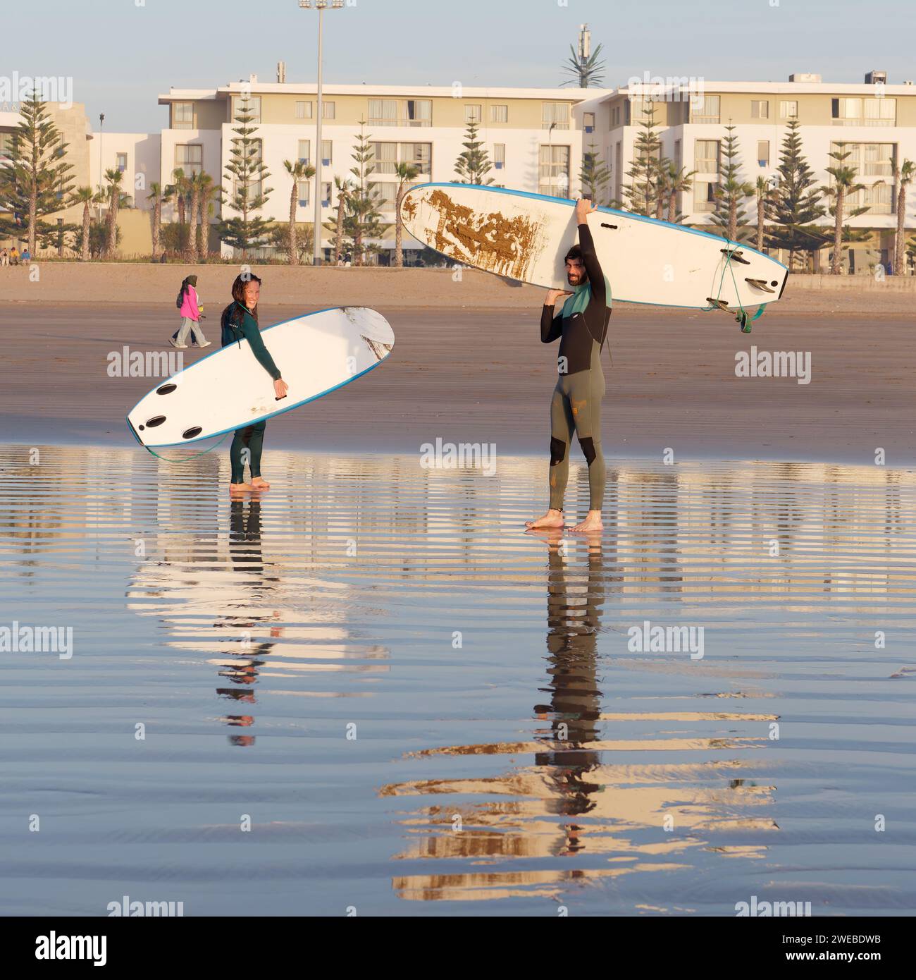 I giovani surfisti sorridono mentre trasportano tavole da surf lontano dal mare su una spiaggia sabbiosa a Essaouira, Marocco, 24 gennaio 2024 Foto Stock