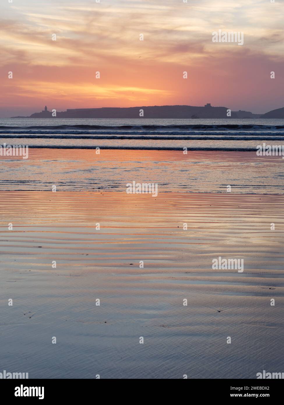 Il tramonto del cielo rosso nuvoloso si riflette nelle acque poco profonde con un'isola alle spalle a Essaouira, Marocco, 24 gennaio 2024 Foto Stock