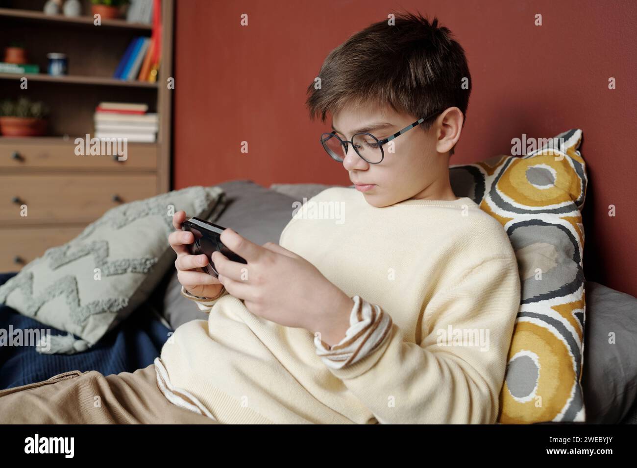 Giovane scolaro con occhiali da vista e pullover seduto sul letto di fronte alla fotocamera e guardando lo schermo dello smartphone mentre si gioca a un videogioco Foto Stock