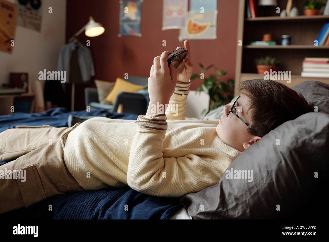 Studente riposante che si rilassa sul letto di fronte alla fotocamera e guarda lo schermo dello smartphone sul gamepad mentre giochi a video game a piacere Foto Stock