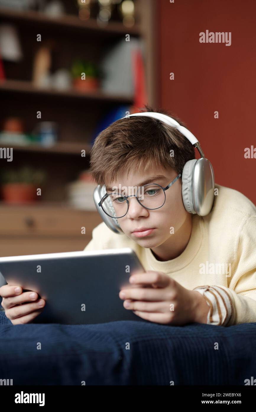Scolaro serio con occhiali da vista e cuffie che guarda lo schermo del tablet mentre guardi film o video musicali online a tuo piacimento Foto Stock