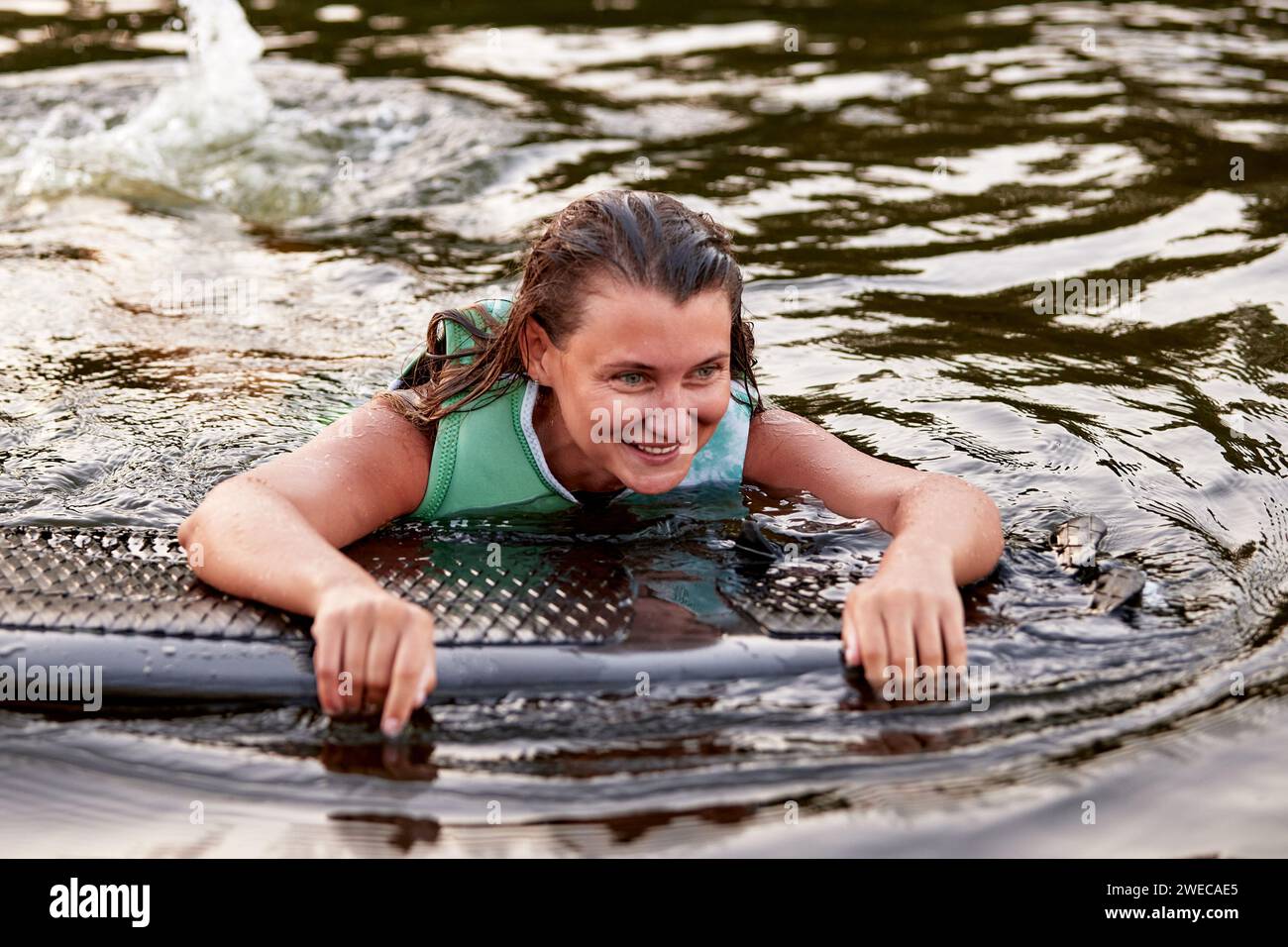 La giovane ragazza sportiva con un giubbotto di salvataggio verde nuota in acqua con un wakeboard in mano. La sportiva felice si sta preparando per un ripido wakebo Foto Stock